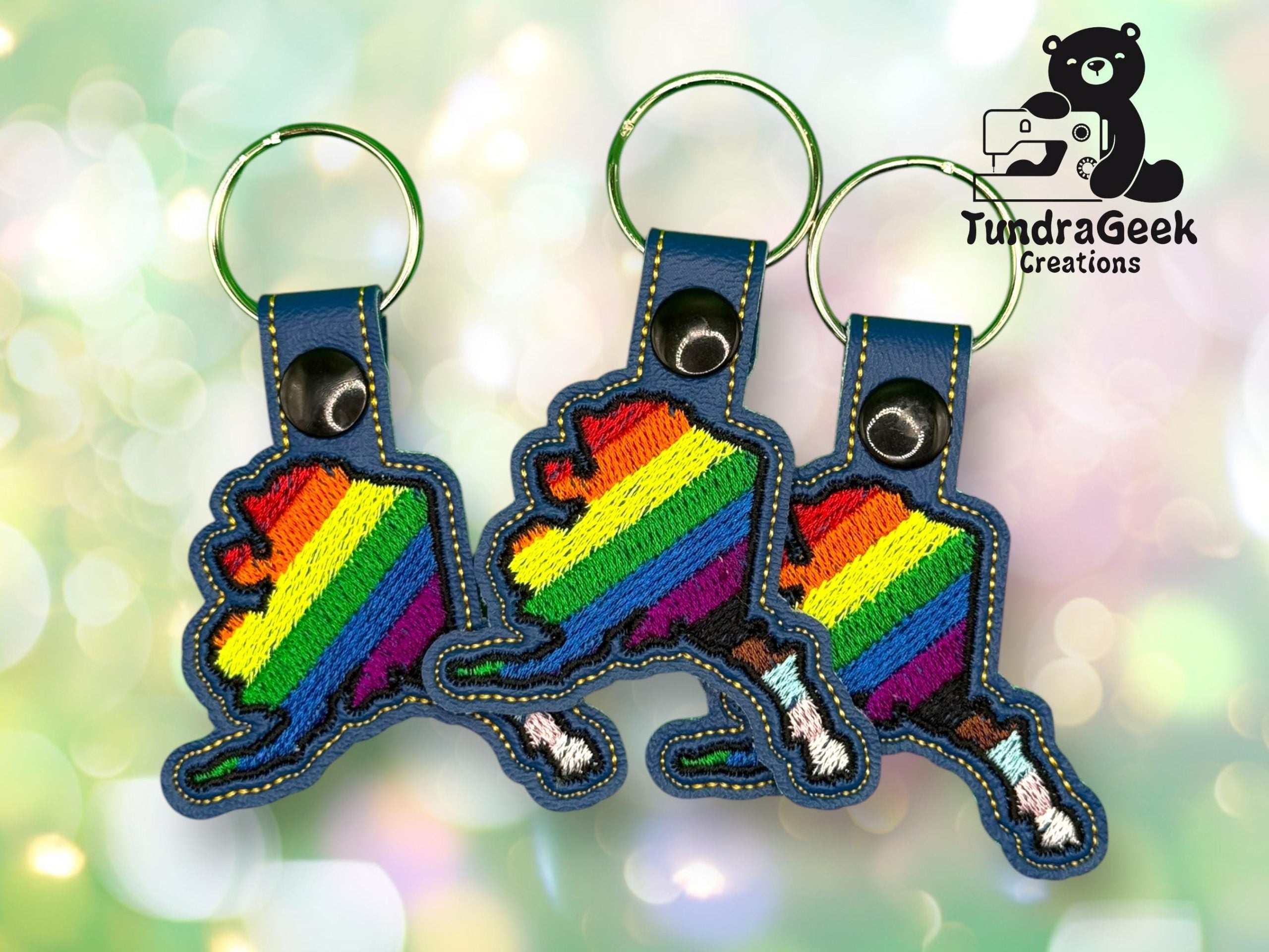 Three blue keychains shaped like Alaska, each featuring rainbow stripes, are displayed with the TundraGeek Creations logo in the top right corner.
