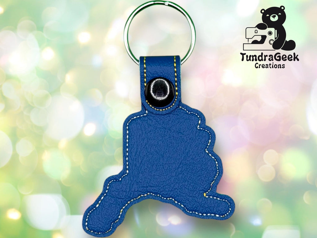 A blue leather keychain in the shape of Alaska with yellow stitching, displayed on a ring, with the TundraGeek Creations logo in the background.