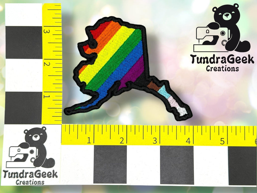 A patch in the shape of Alaska with a rainbow pride design is placed on a measurement grid with TundraGeek Creations logos in the image corners.