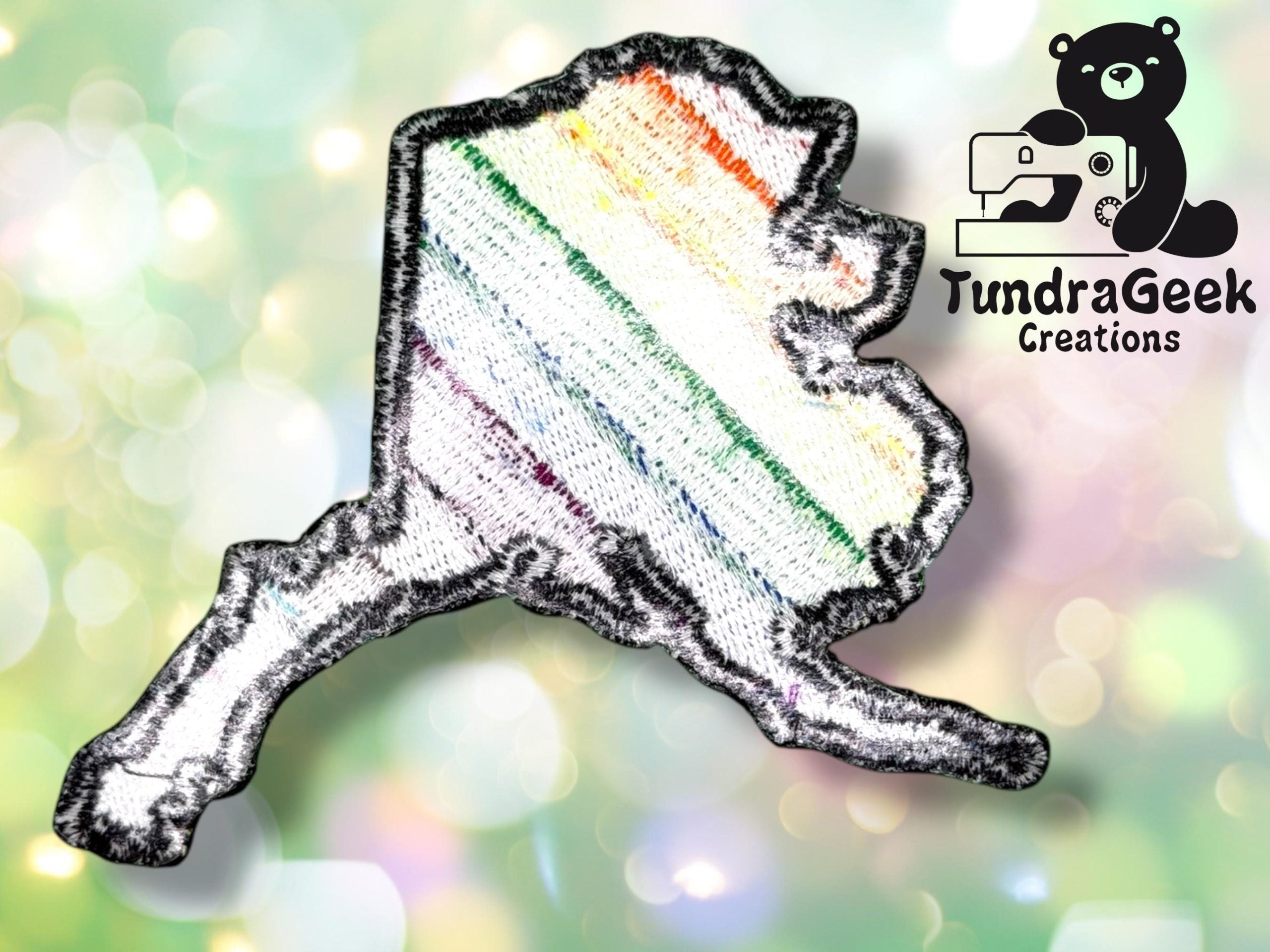 A patch in the shape of Alaska with diagonal rainbow stripes, shown on a blurred background. TundraGeek Creations logo with a bear and a sewing machine is in the upper right corner.