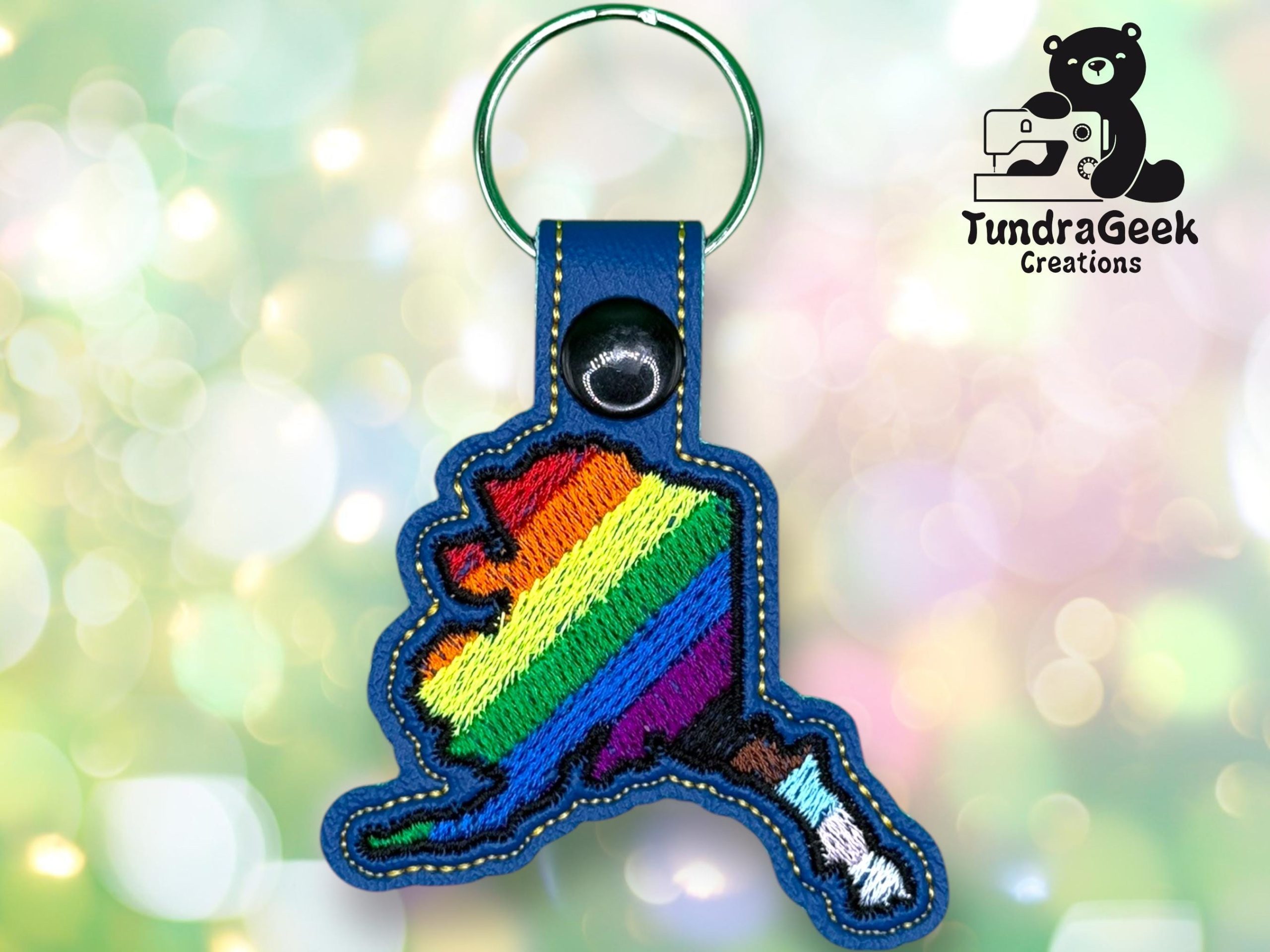 A keychain shaped like Alaska, embroidered with rainbow stripes, is displayed on a blurred colorful background. TundraGeek Creations logo appears in the top right corner.