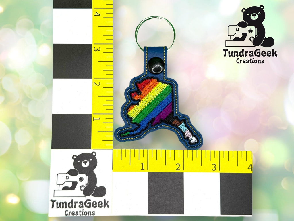 A keychain in the shape of Alaska with rainbow stripes is placed beside a ruler for scale. The TundraGeek Creations logo appears in two corners.