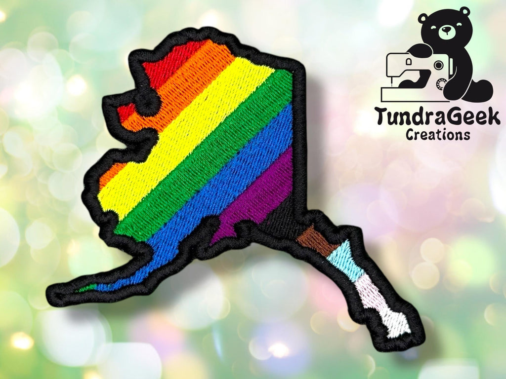 Embroidered patch in the shape of Alaska with rainbow pride stripes, featuring the TundraGeek Creations logo with a sewing bear in the corner.