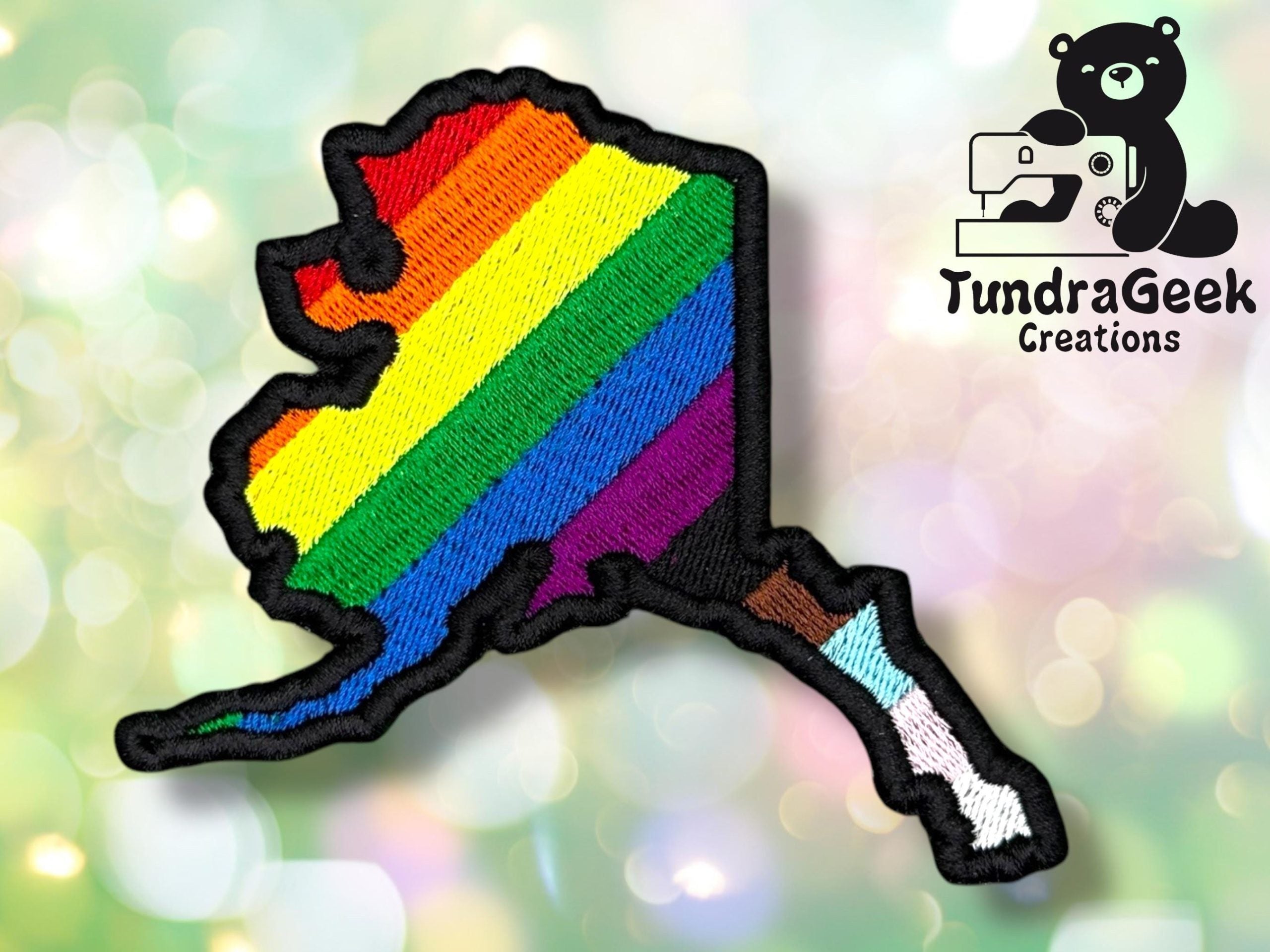 Embroidered patch in the shape of Alaska with rainbow pride stripes, featuring the TundraGeek Creations logo with a sewing bear in the corner.
