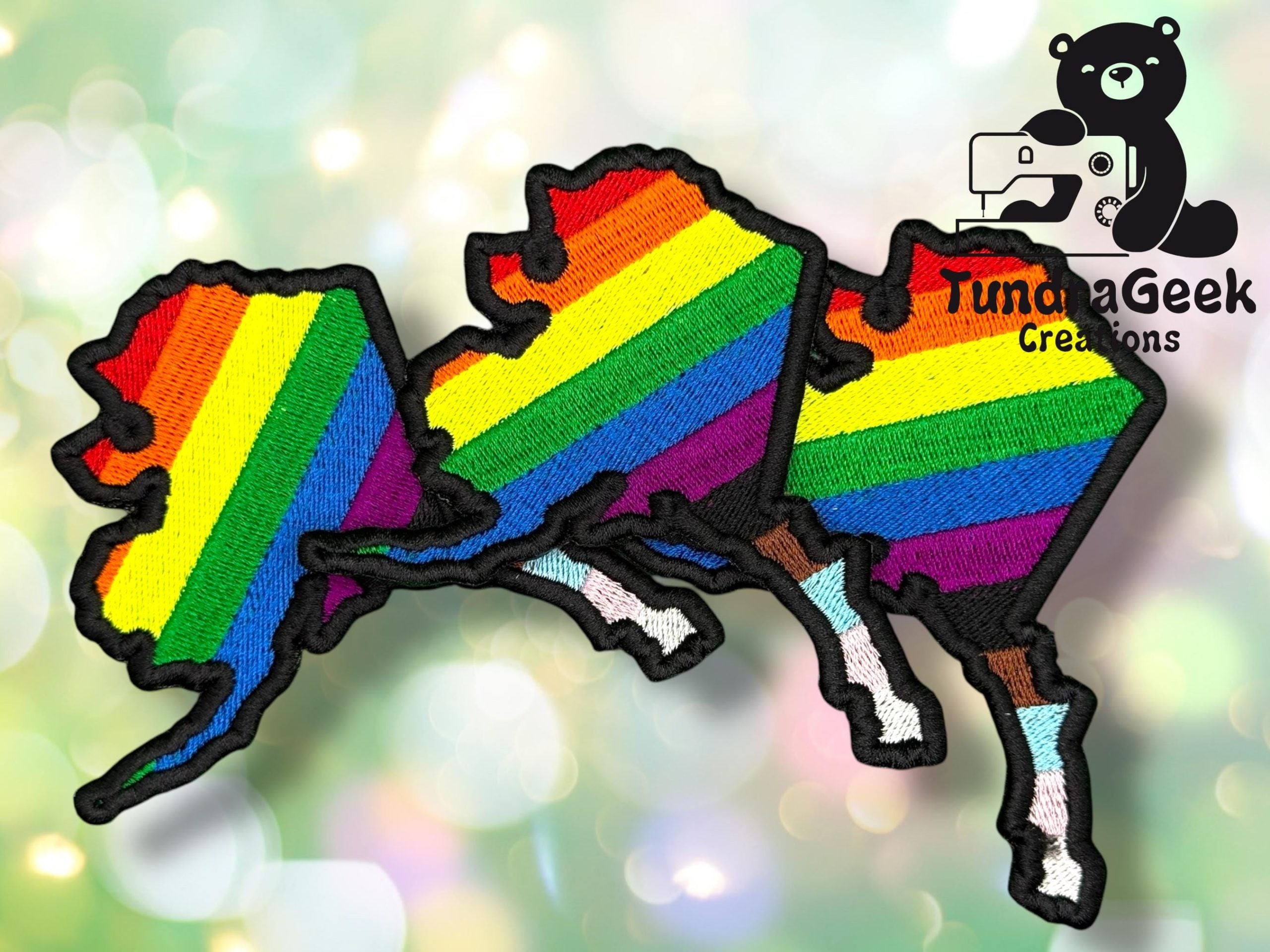 Three embroidered patches in the shape of Alaska feature rainbow pride stripes. The logo TundraGeek Creations with a bear holding a camera appears in the corner.