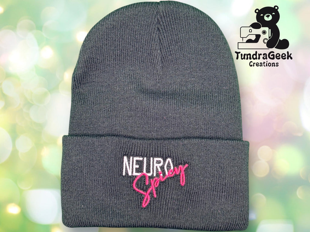 A gray knit beanie with the words NEURO Spicy embroidered in white and pink. The TundraGeek Creations logo with a bear and sewing machine is in the top right corner.