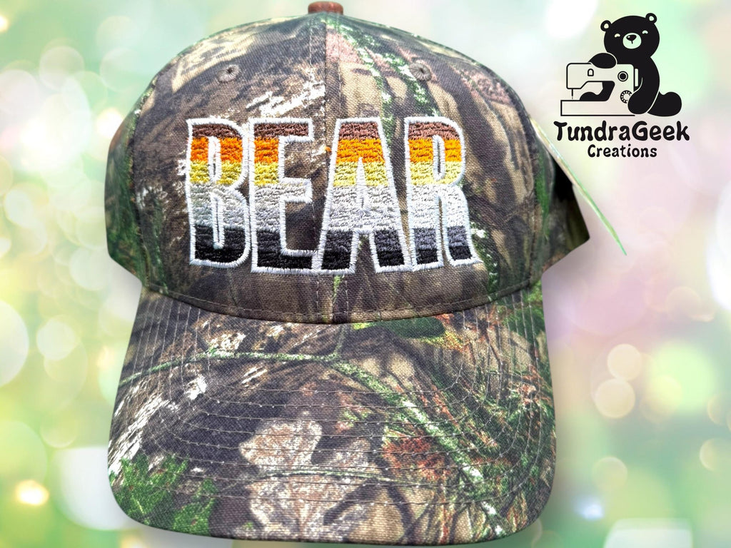 A camouflage-patterned cap with BEAR embroidered in bold, gradient letters; TundraGeek Creations logo with a bear and sewing machine is in the top right corner.