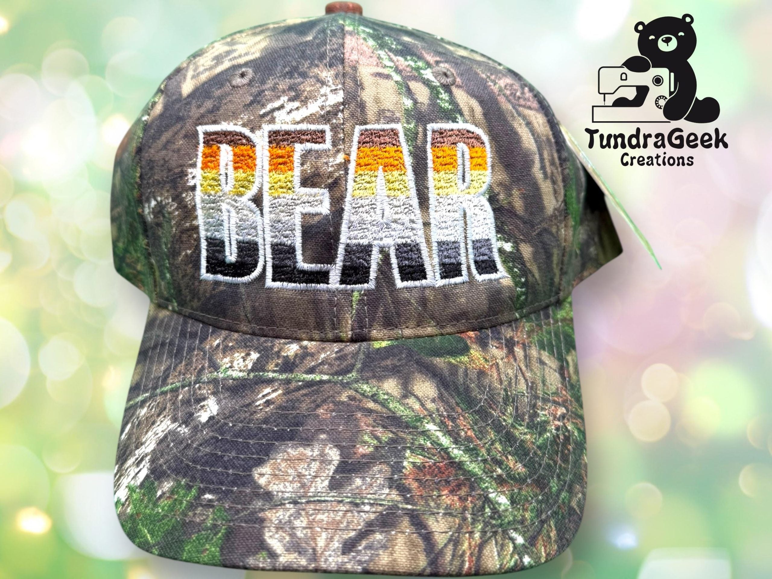 Camouflage cap with the word BEAR embroidered in large letters with a gradient color effect; TundraGeek Creations logo featuring a bear and sewing machine is in the top right corner.