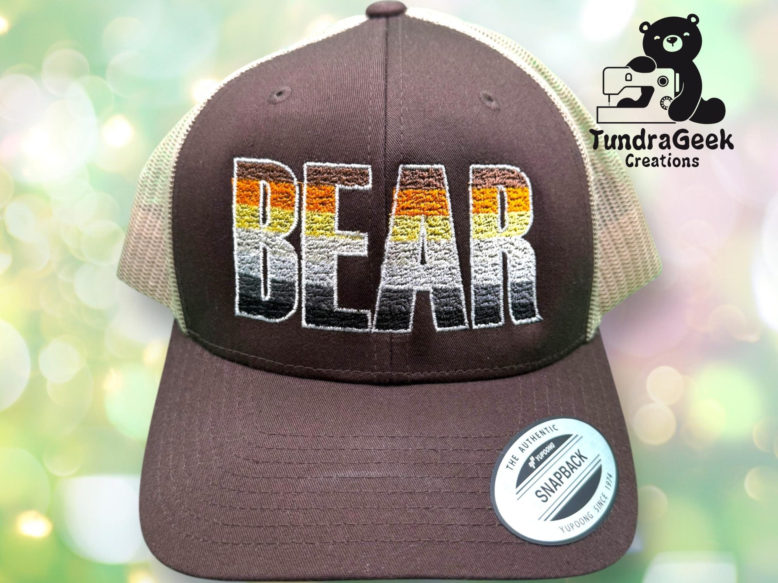 A trucker hat with the word BEAR embroidered in brown, orange, and white stripes, with a Snapback sticker on the brim and TundraGeek Creations logo in the background.