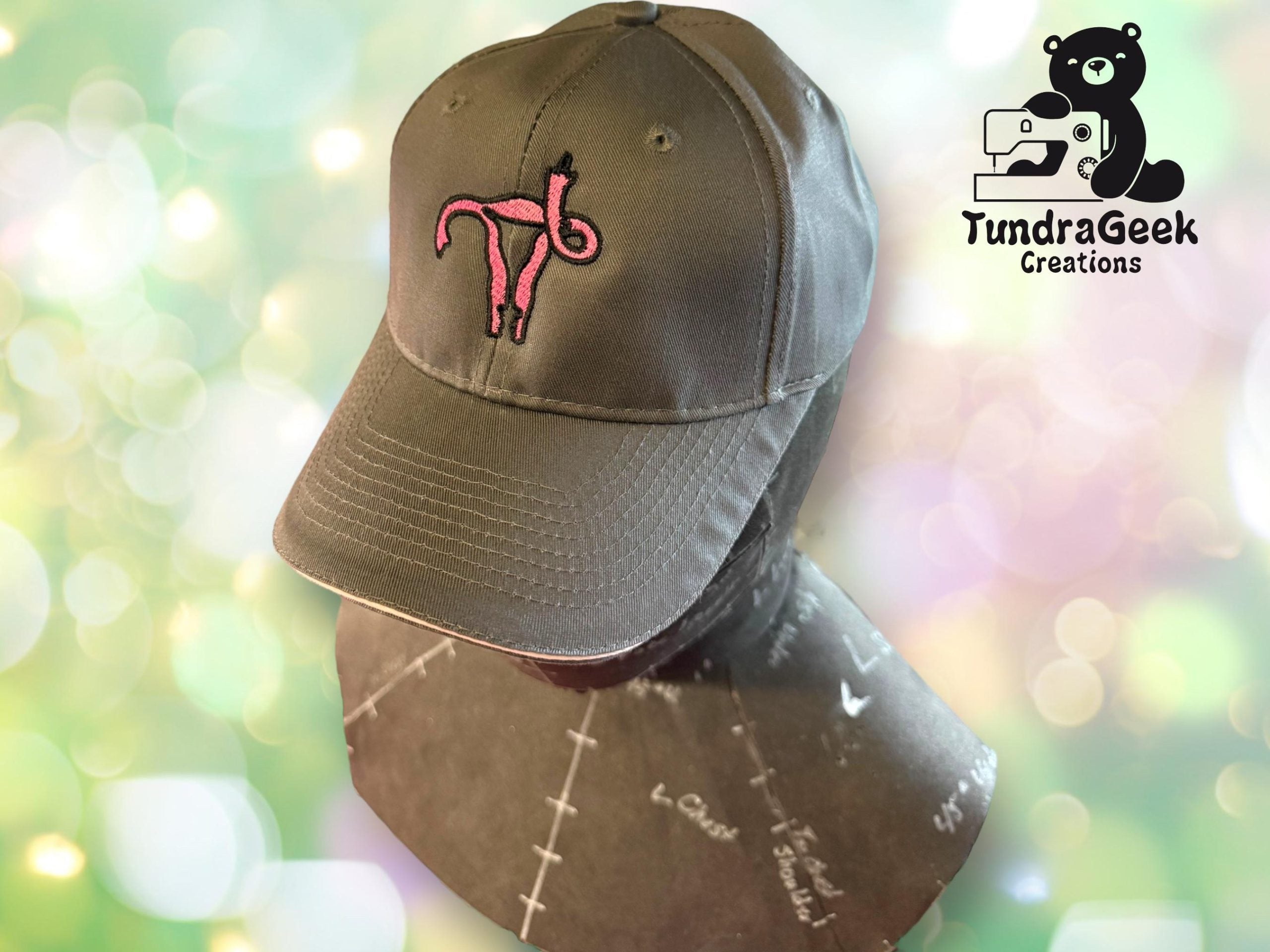 Gray baseball cap with a pink uterus design on the front, displayed on a mannequin head. The TundraGeek Creations logo appears in the top right corner.