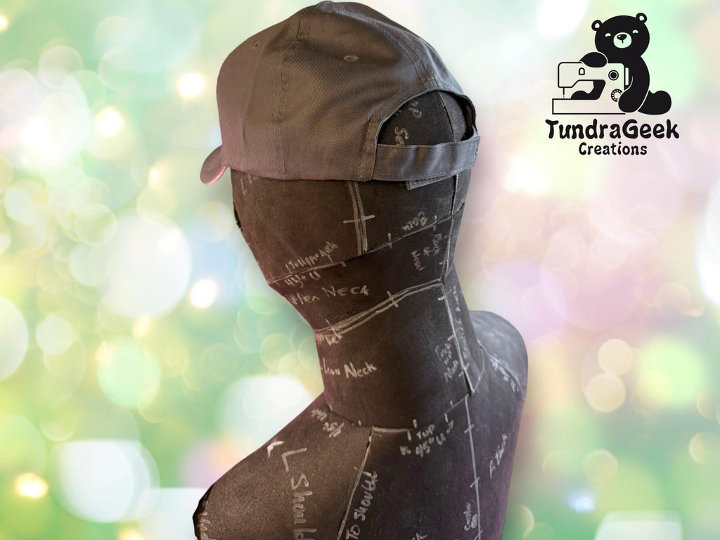 A gray baseball cap on a black mannequin with pattern markings, set against a blurred green background. TundraGeek Creations logo is in the top right corner.