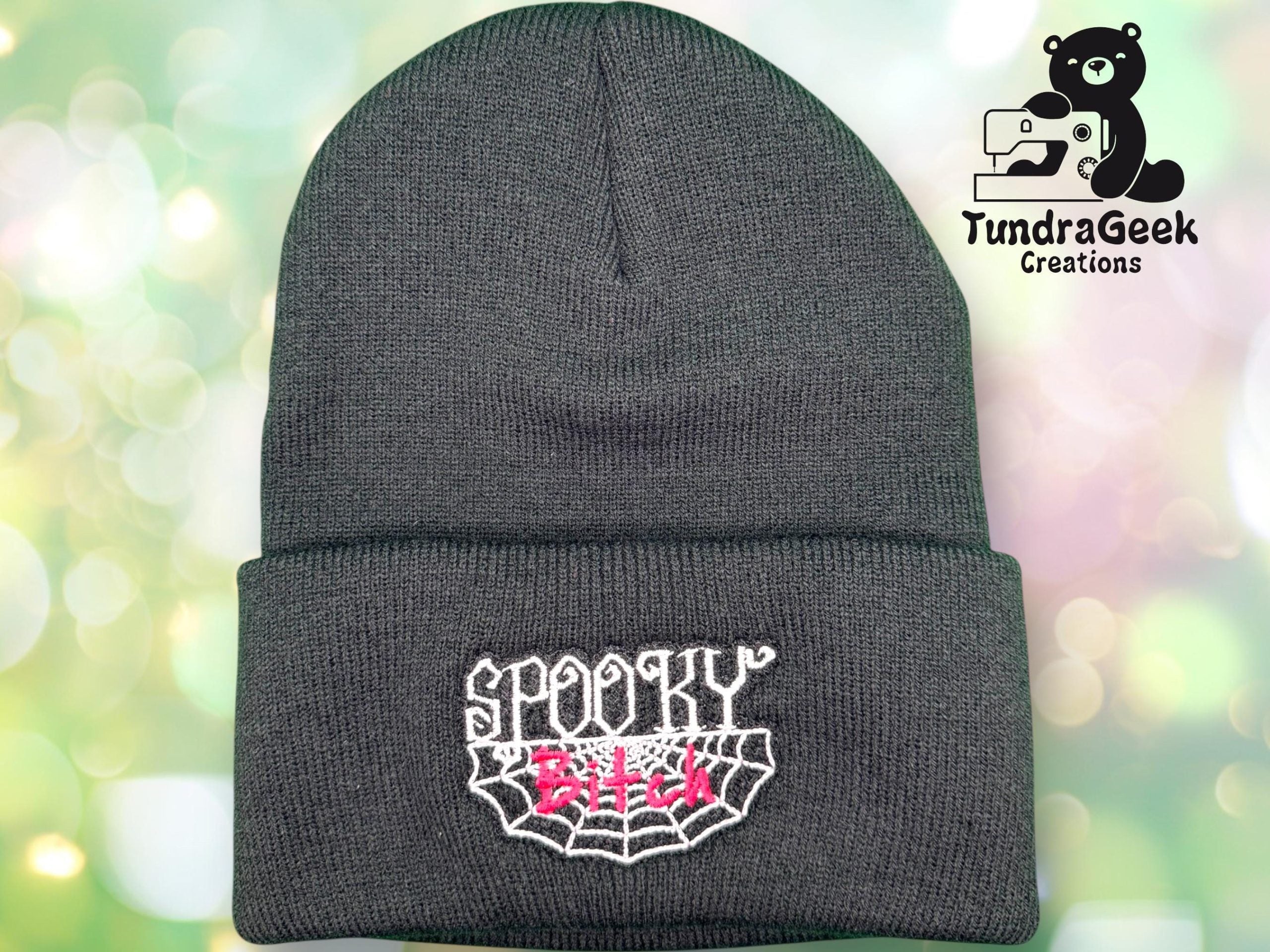 A dark knit beanie with Spooky Bitch embroidered in white and pink above a spider web design. The TundraGeek Creations logo appears in the top right corner.