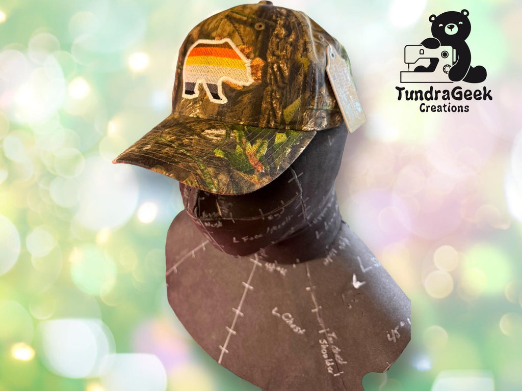 Camo baseball cap with a bear patch featuring rainbow stripes, displayed on a dress form. Background shows the TundraGeek Creations logo with a bear and sewing machine.