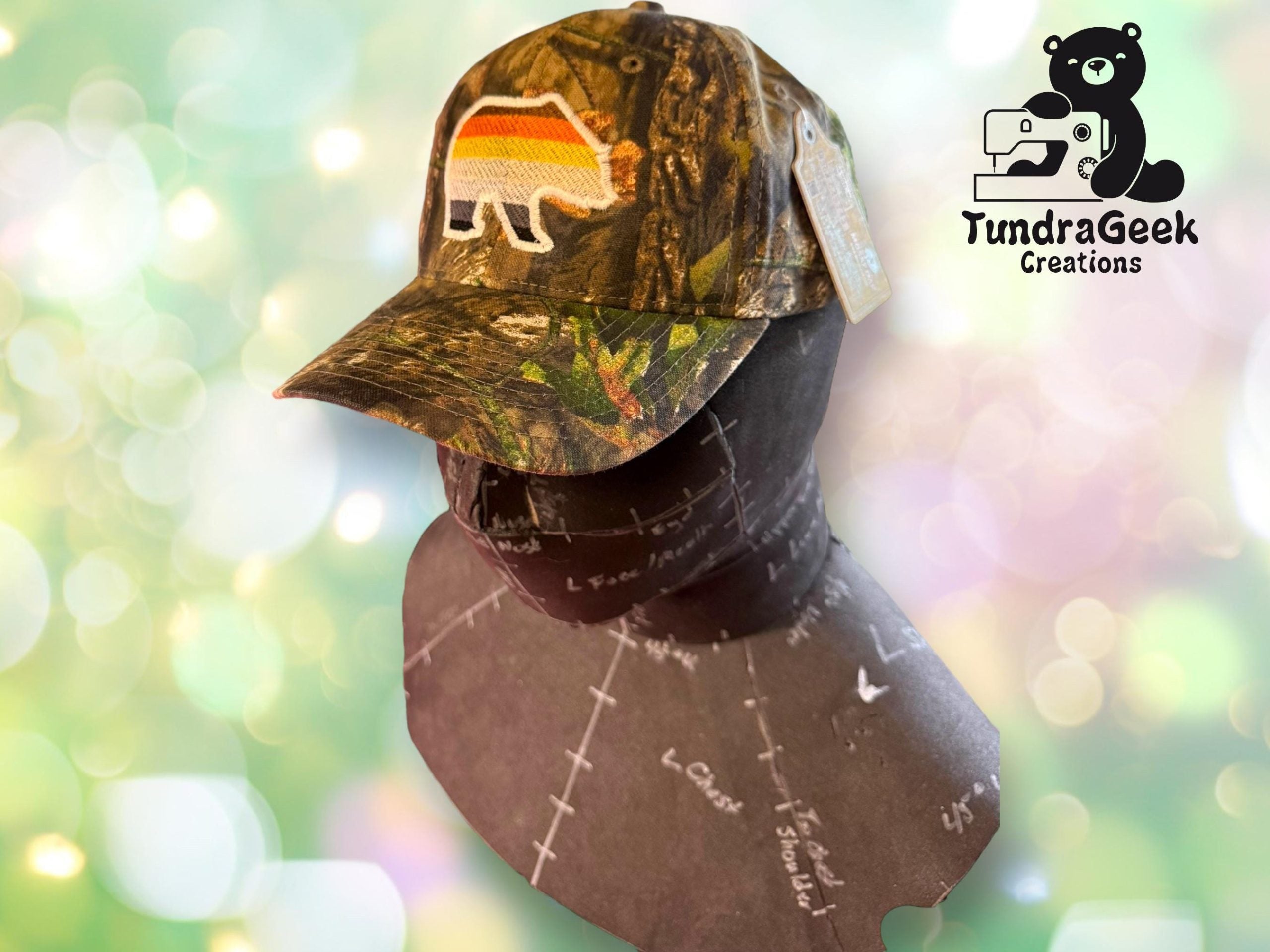 Camo baseball cap with a bear patch featuring rainbow stripes, displayed on a dress form. Background shows the TundraGeek Creations logo with a bear and sewing machine.