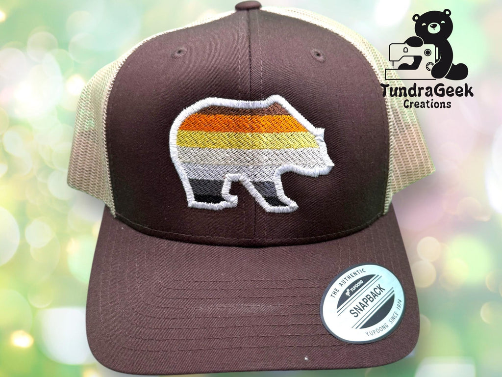 Brown and tan mesh trucker hat with an embroidered bear featuring horizontal stripes in yellow, orange, and gray. TundraGeek Creations logo with a bear and sewing machine is in the background.