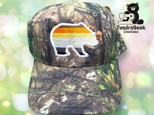 A camouflage baseball cap with an embroidered bear outline filled with brown, orange, yellow, and white stripes. TundraGeek Creations logo is in the top right corner.