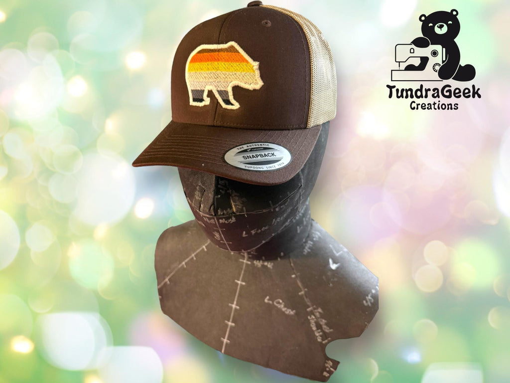 A brown and beige trucker hat with a retro bear patch is displayed on a sewing mannequin, with the TundraGeek Creations logo in the background.