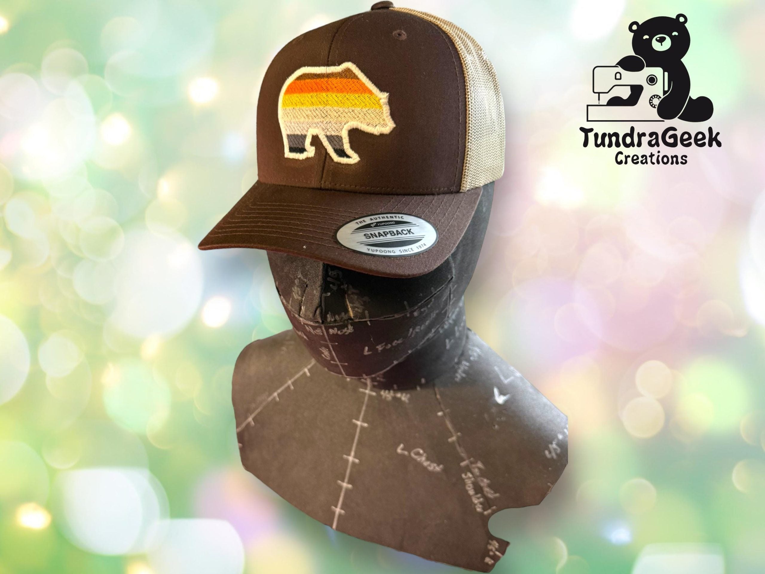 A brown and beige trucker hat with a retro bear patch is displayed on a sewing mannequin, with the TundraGeek Creations logo in the background.