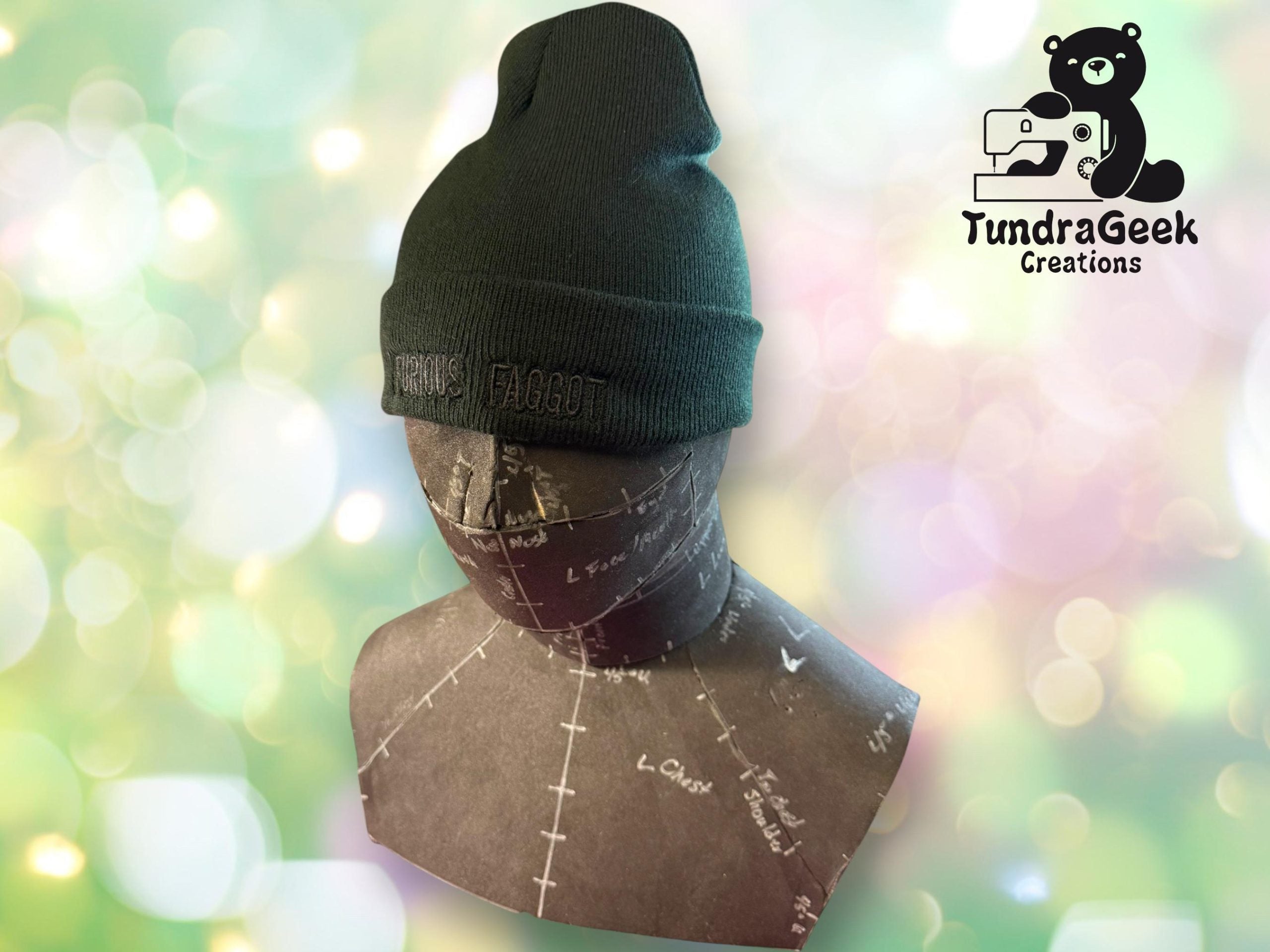 A mannequin head wearing a dark beanie with embroidered text, displayed in front of a light, bokeh background with the TundraGeek Creations logo in the corner.
