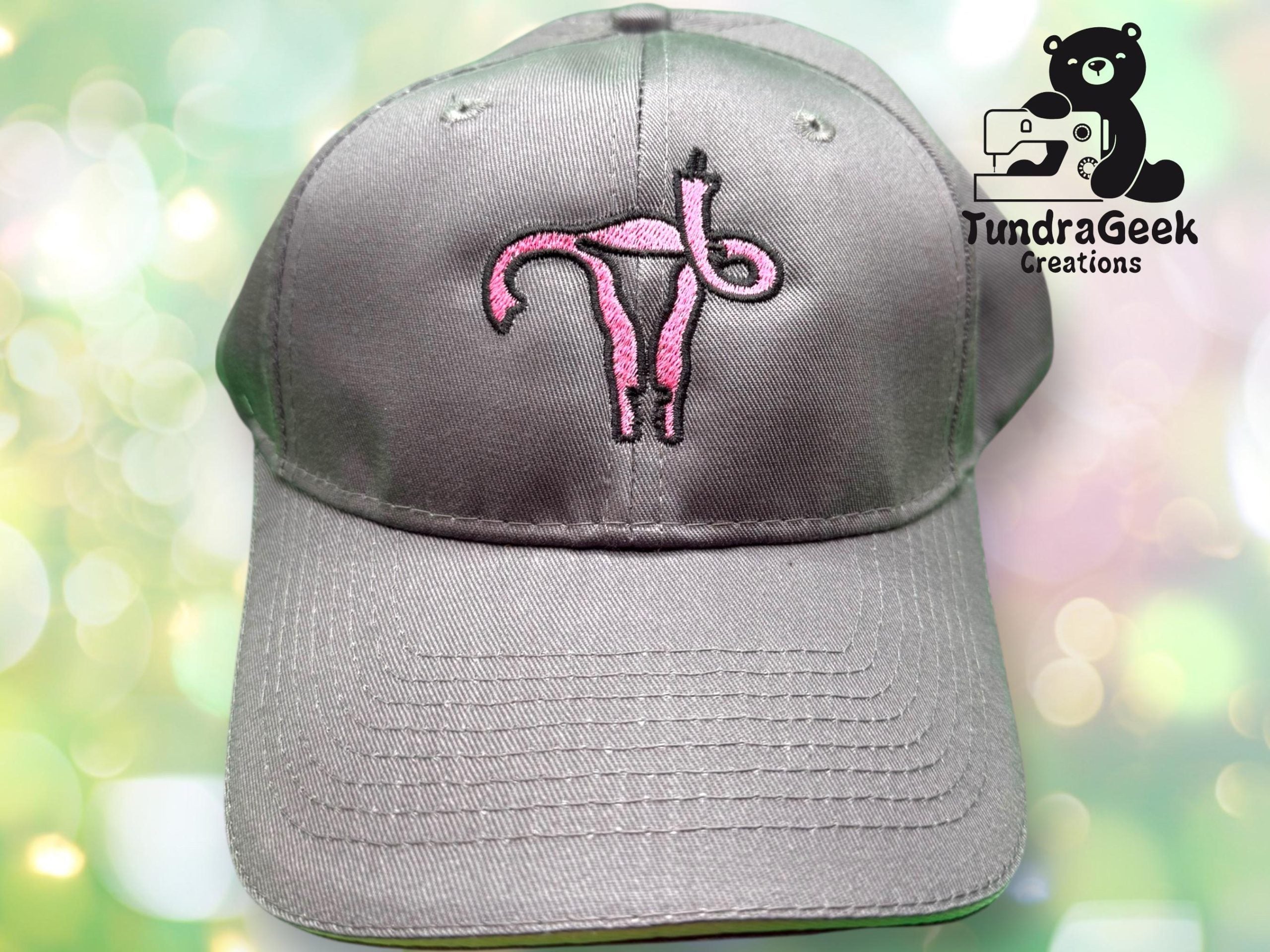 Gray baseball cap featuring a pink embroidered uterus design on the front. TundraGeek Creations logo with a bear and sewing machine appears in the upper right corner.