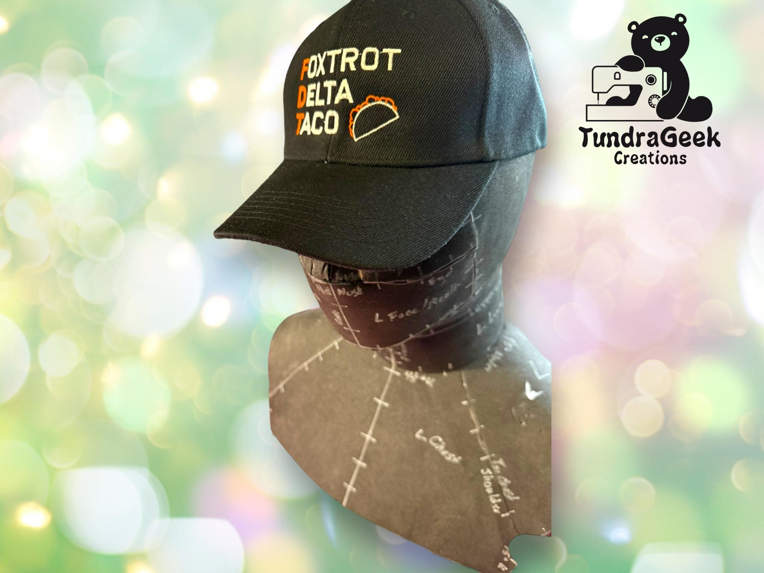 Mannequin head wearing a black Foxtrot Delta Taco hat with a taco graphic, set against a blurred background with TundraGeek Creations logo in the corner.