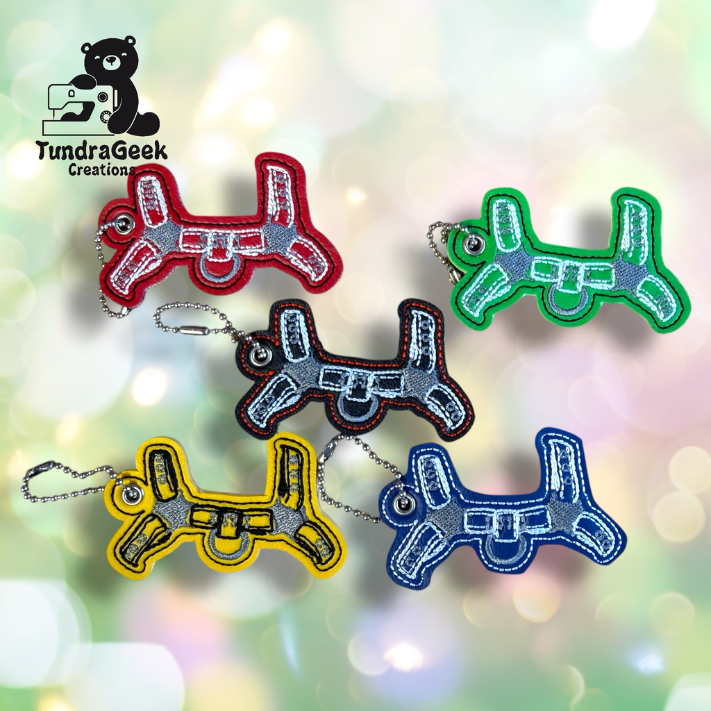 Six embroidered keychains shaped like soldering irons in red, green, yellow, blue, black, and gray, are displayed on a blurred pastel background with the TundraGeek Creations logo.