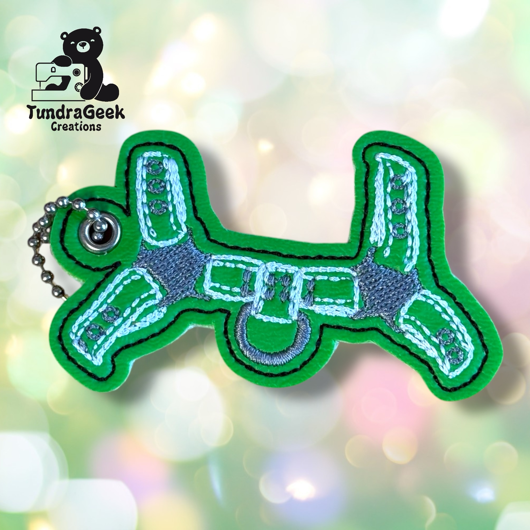 Keychain shaped like a green balloon dog with embroidered details, shown against a blurred light background. TundraGeek Creations logo is in the upper left corner.