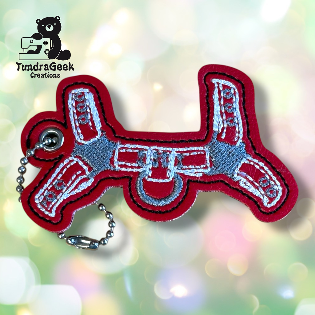 Red and gray embroidered keychain shaped like a balloon dog, with a ball chain attached, on a blurred, light-colored background. TundraGeek Creations logo in the top left corner.
