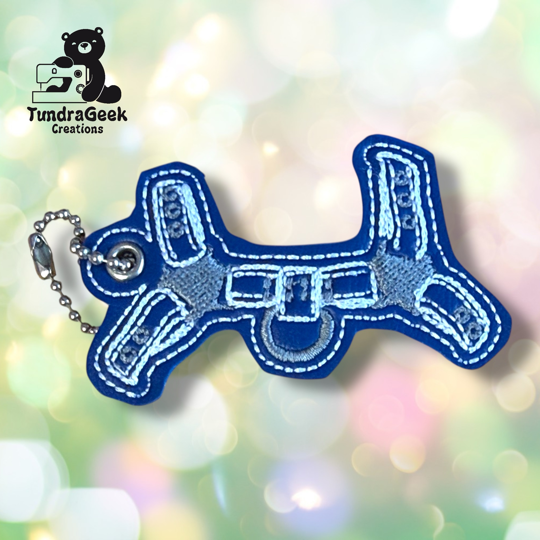Blue and gray airplane-shaped keychain with white stitching, displayed on a blurred pastel background. TundraGeek Creations logo is visible in the top left corner.