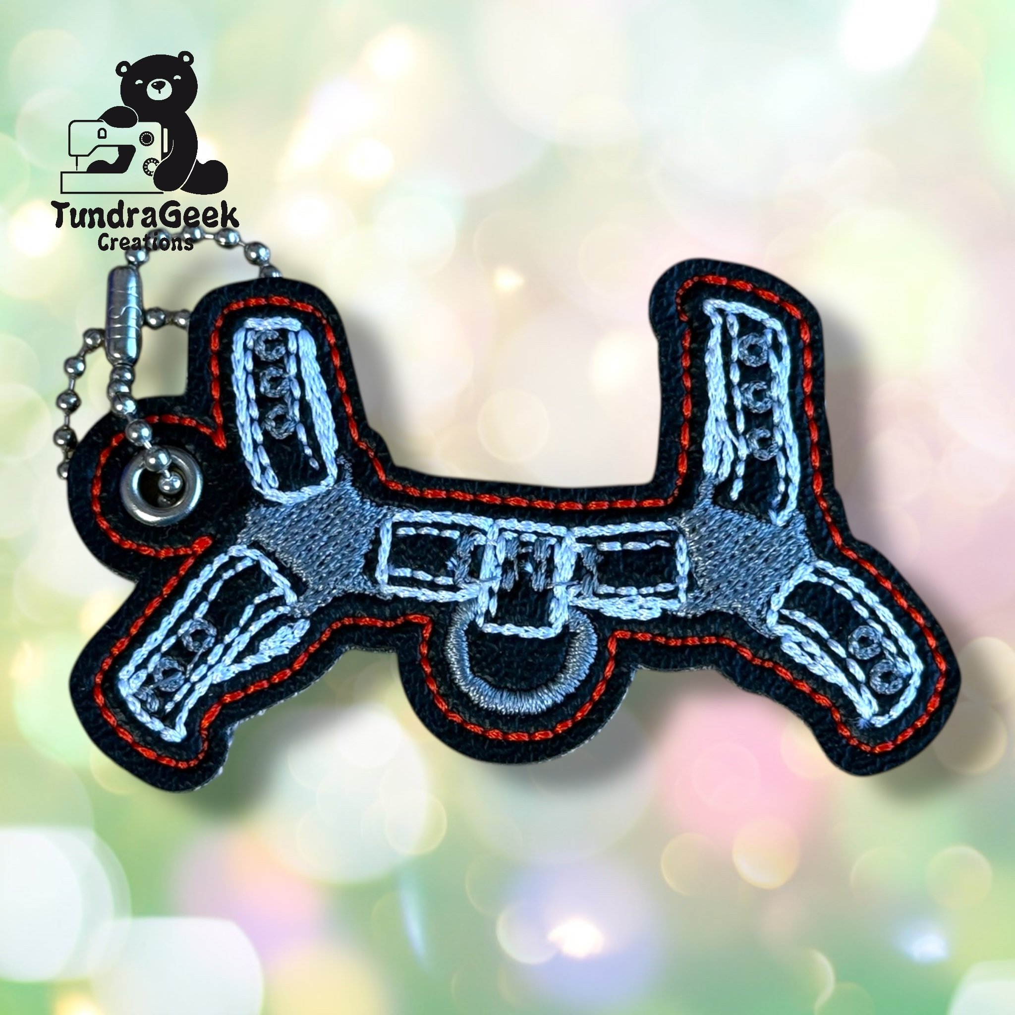 Keychain shaped like a drone with embroidered gray, white, and orange details, attached to a ball chain. The TundraGeek Creations logo is visible in the background.