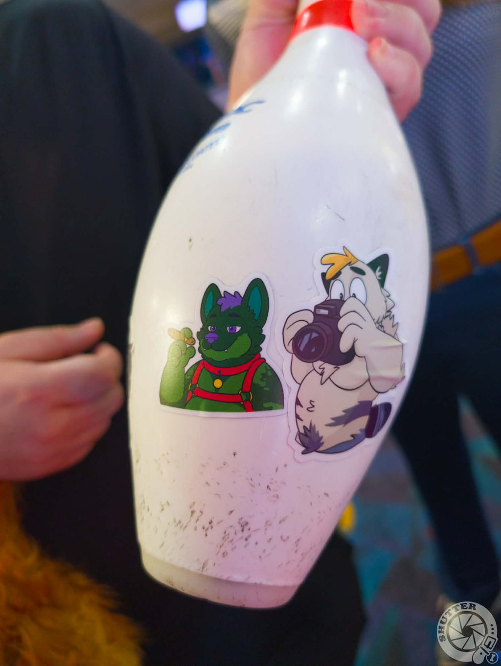 A white bowling pin with two anthro character stickers presented by someone's hand. Photo taken by Shutterbear (Bluesky)