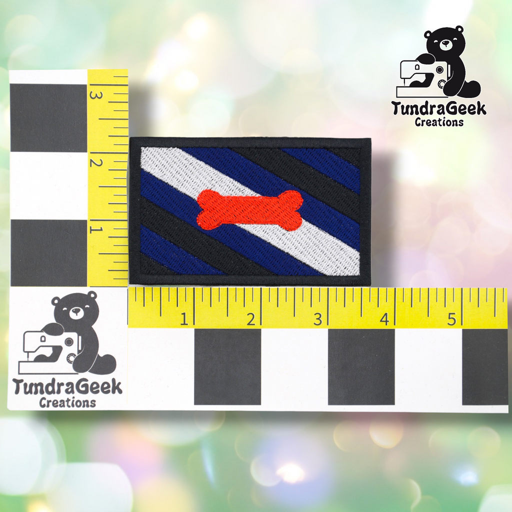 The Pup Pride Flag Patch features diagonal black and blue stripes, a white stripe, and a red bone, shown on a measurement grid with TundraGeek Creations logos. Perfect embroidered sew-on or iron-on patch for pup play gear!.